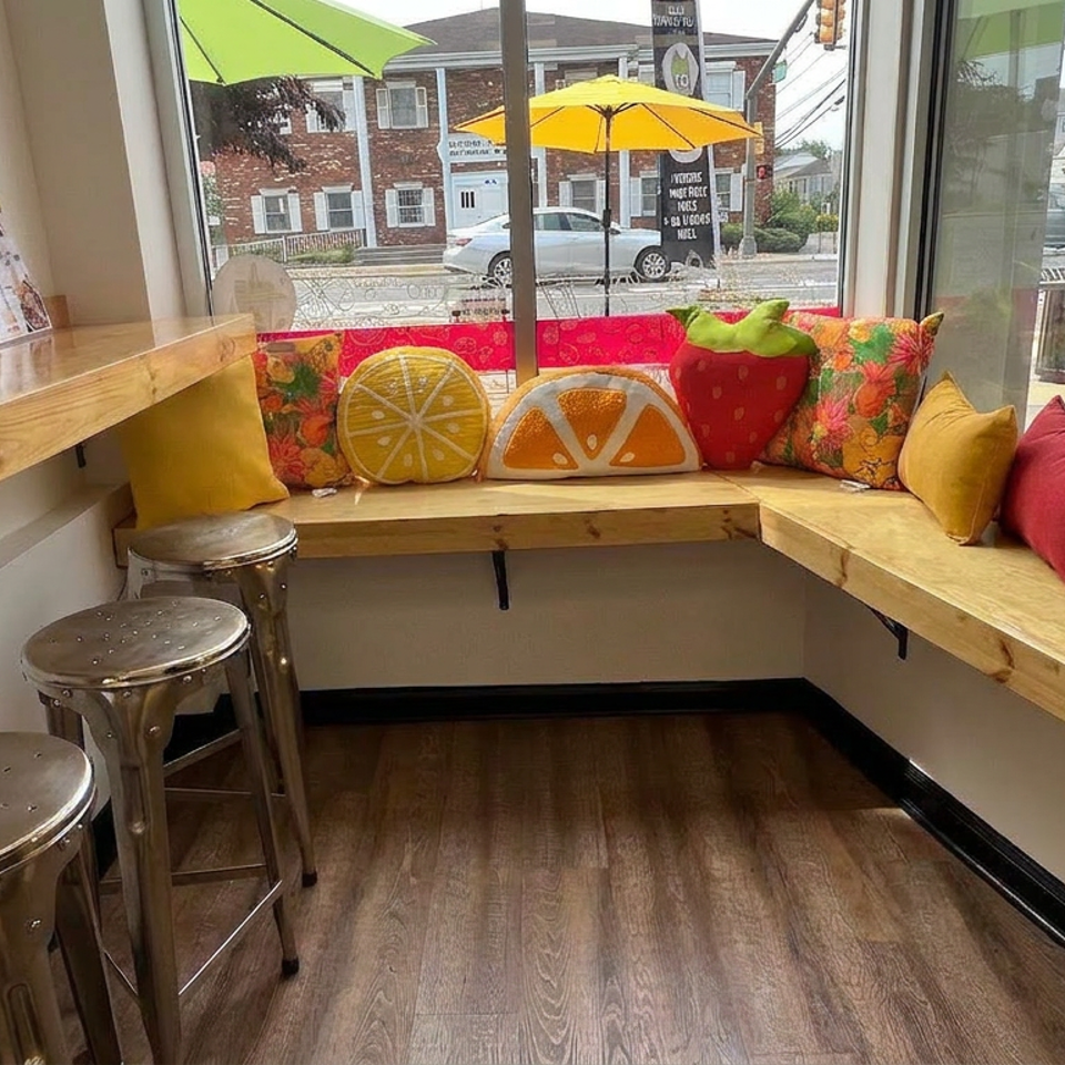 Bright restaurant seating area with wooden counters, metal stools, colorful fruit-shaped pillows, and large street-facing windows.