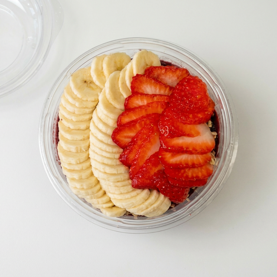 Clear bowl of sliced bananas and strawberries arranged over a layered dessert, with its lid beside it.
